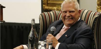 Doug Ford sitting in a chair, holding a microphone and smiling while listening to someone. 