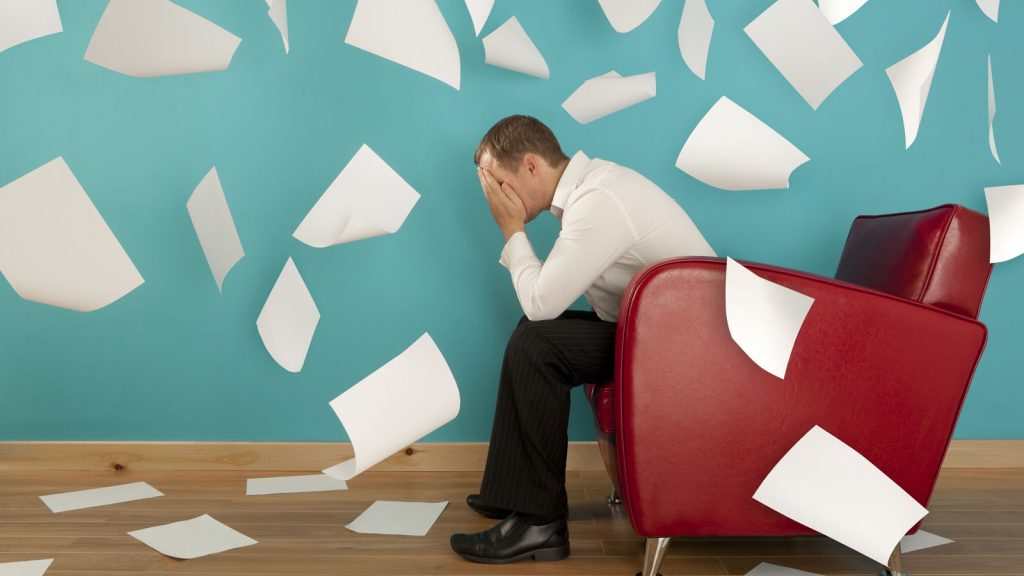 Man sitting in an arm chair, head in his hands. Around him, sheets of paper fall around him and on the floor.