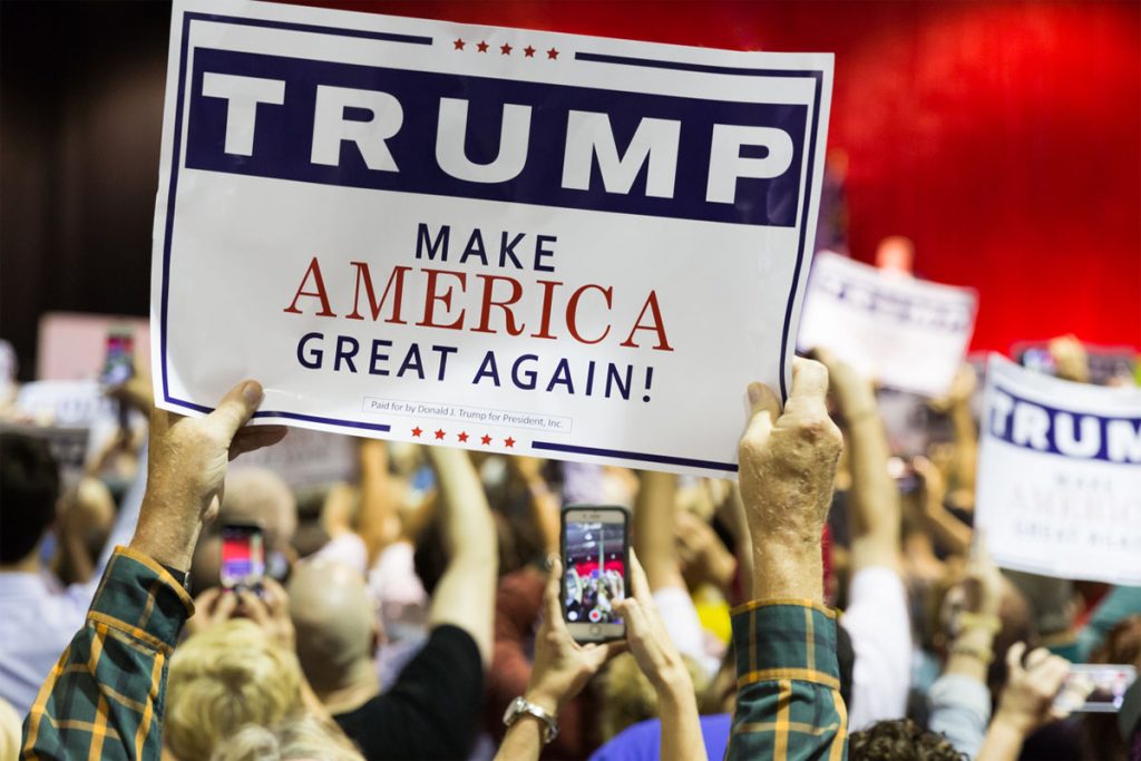 A large crowd. Arms in the air hold up a sign that says "Trump: Make America Great Again."